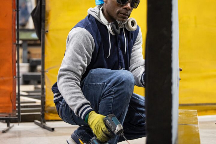 A man wearing sunglasses, a blue bandana, and gloves crouches indoors in Fires, Ohio, using a cordless drill on a piece of wood, with yellow panels in the background.