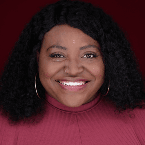 A woman with curly black hair, large hoop earrings, and a nose ring smiles at the camera. She is wearing a burgundy top against a matching burgundy background.
