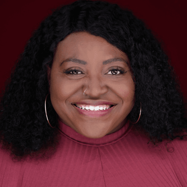 A woman with curly black hair, large hoop earrings, and a nose ring smiles at the camera. She is wearing a burgundy top against a matching burgundy background.