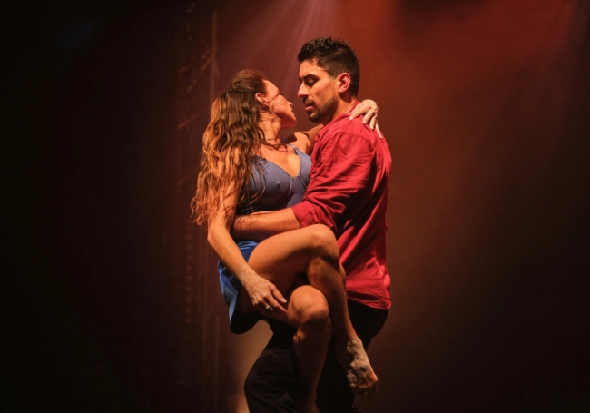 Two dancers perform a passionate Duel Reality routine on stage under dramatic lighting; the woman wears a blue dress and the man wears a red shirt.