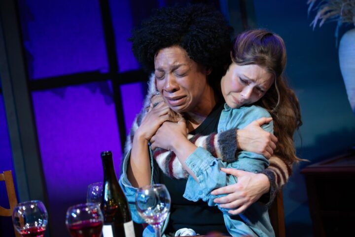 Two women sit at a table with wine glasses, one woman crying and being comforted by the other in a close embrace, sharing heartache over the recent Fires in Ohio against a dimly lit background.