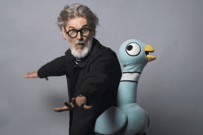 A man with gray hair and glasses poses with his arms outstretched, standing next to a large blue bird puppet against a plain background—evoking the playful spirit of Naked Mole Rat Gets Dressed.