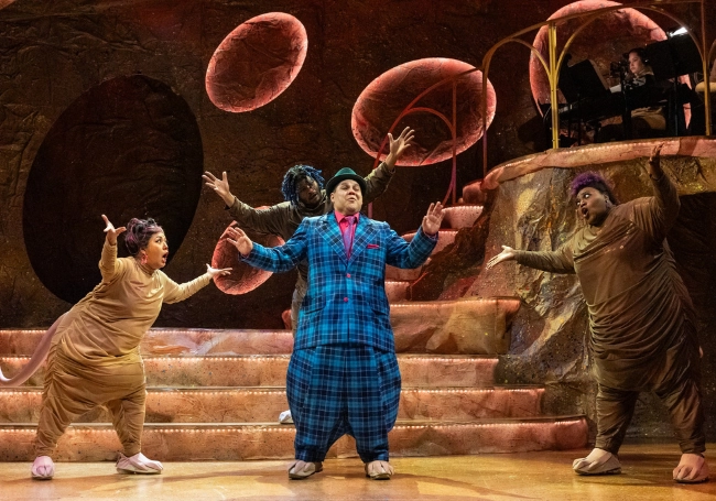Four performers in animal costumes and one in a blue plaid suit—just like Wilbur from Naked Mole Rat Gets Dressed—sing and gesture dramatically on a stage with rounded, earth-toned set pieces and a musician visible in the background.