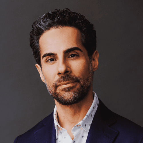 A man with short dark hair and a trimmed beard, wearing a dark suit jacket and a patterned white shirt, poses against a plain dark background.