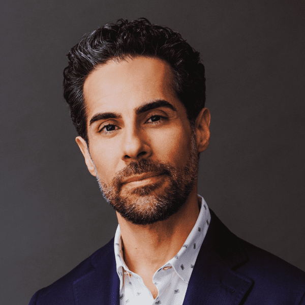 A man with short dark hair and a trimmed beard, wearing a dark suit jacket and a patterned white shirt, poses against a plain dark background.