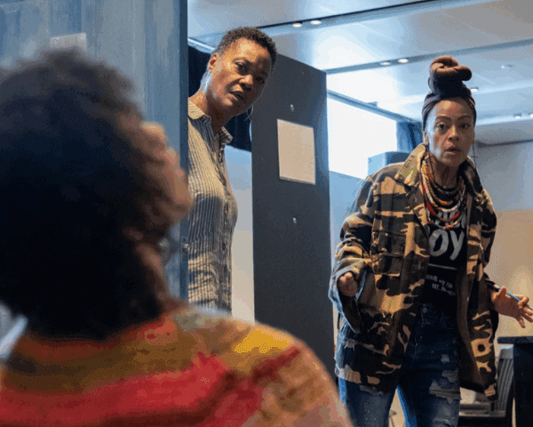 Three women interact indoors; one sits in the foreground, while two others stand behind a door, engaged in conversation or a dramatic moment.