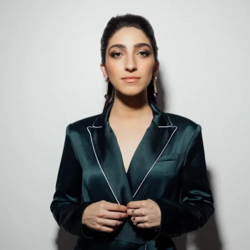Emily Estefan with long dark hair in a ponytail, wearing a dark green satin suit with white trim, stands against a plain light background and looks directly at the camera.