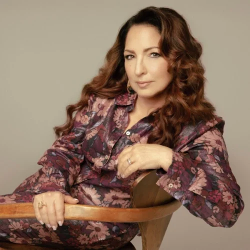 A woman with long, wavy brown hair sits on a wooden chair in a floral-patterned outfit, gazing at the camera with a neutral expression, reminiscent of Gloria Estefan's effortless style.