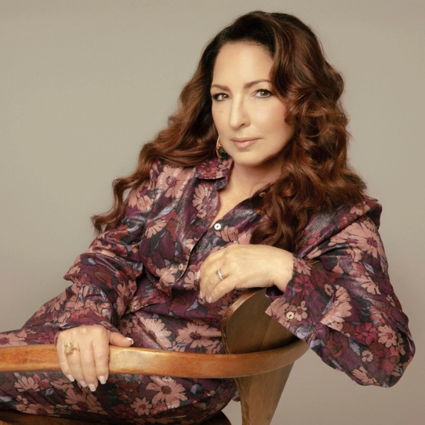 A woman with long, wavy brown hair sits on a wooden chair in a floral-patterned outfit, gazing at the camera with a neutral expression, reminiscent of Gloria Estefan's effortless style.