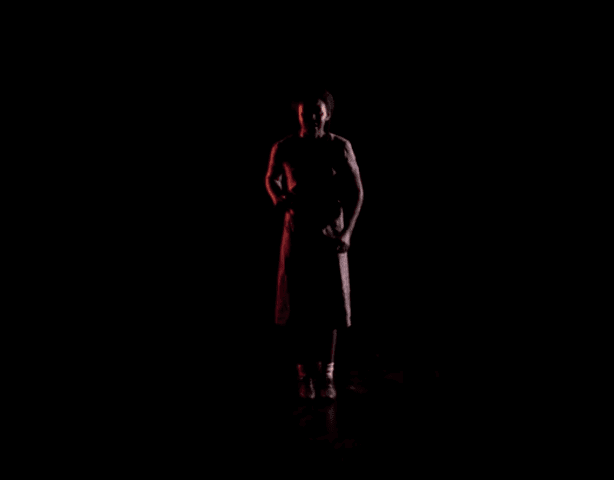 A person stands alone in dim lighting, centered against a black background. Their figure is partially illuminated, highlighting their face and upper body while the rest fades into shadow.