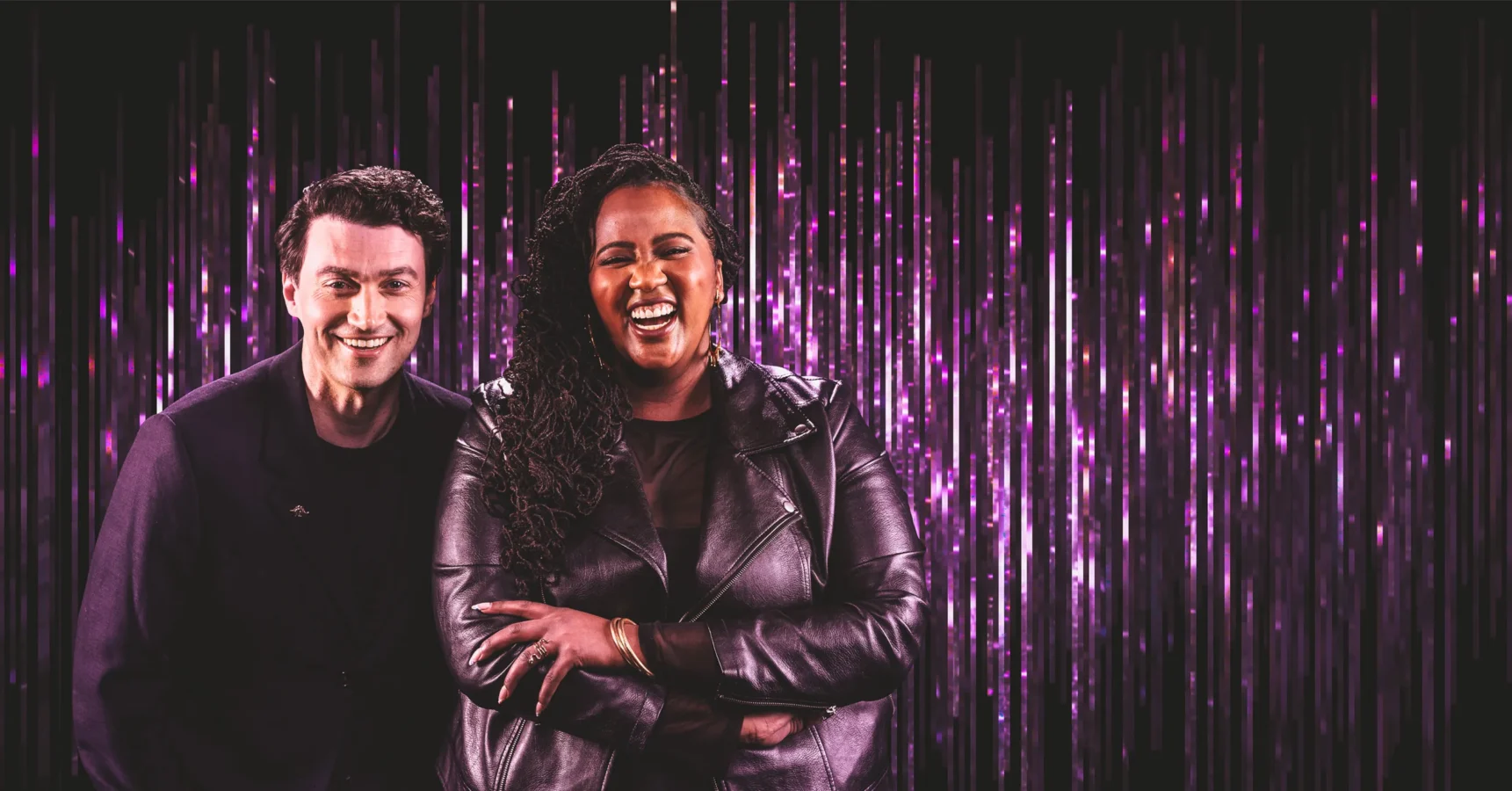 Two people stand side by side, smiling and laughing, in front of a backdrop of vertical purple light streaks.