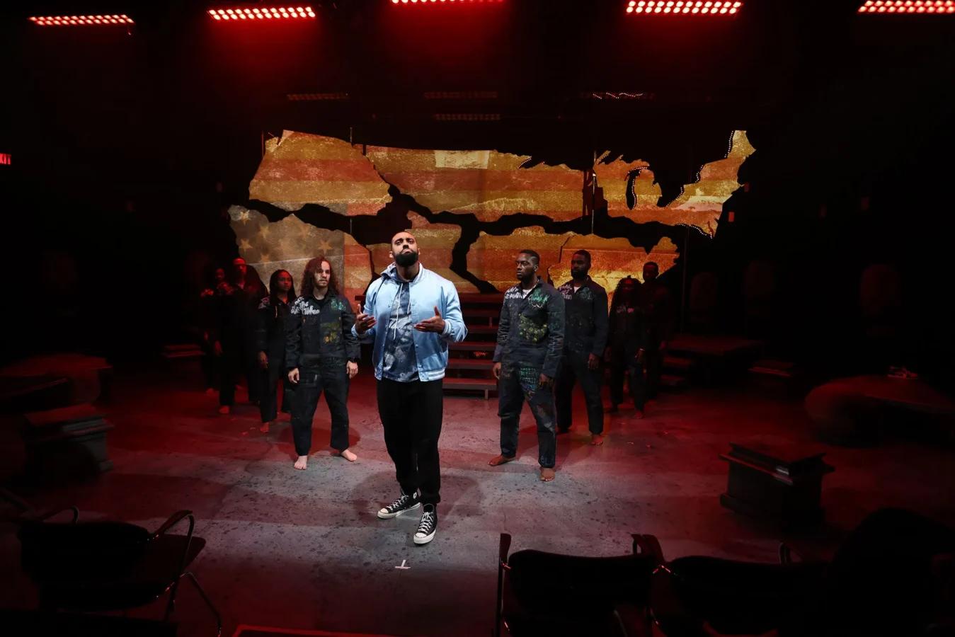 A group of actors stands on a stage with a map of the United States projected behind them; one person stands at the front, singing or speaking.