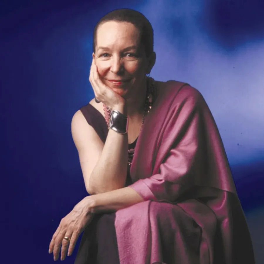 A person with short hair, wearing a sleeveless top and a pink shawl, sits with one hand resting on their face against a blue background.