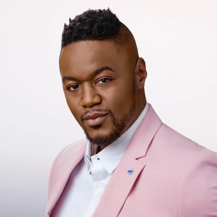 Man with short hair and trimmed beard wearing a light pink suit jacket over a white shirt, facing the camera with a neutral expression against a plain background.