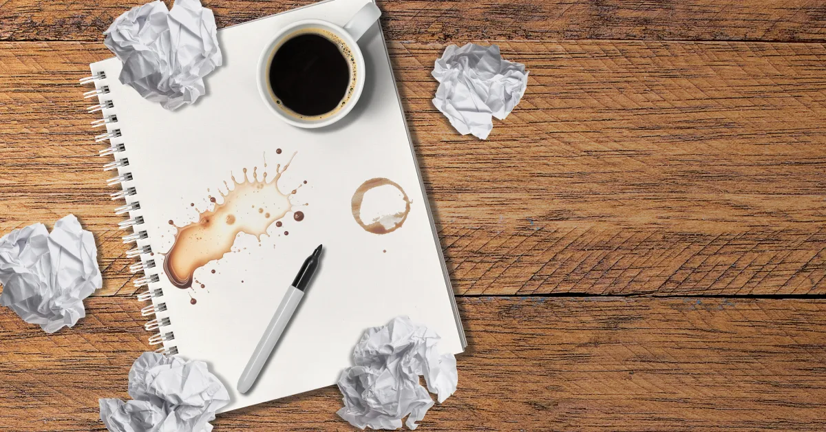 A notebook with coffee stains, a pen, and a cup of coffee on a wooden surface, surrounded by crumpled paper balls.