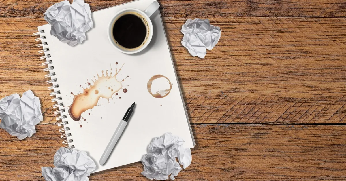 A notebook with coffee stains, a black pen, and a cup of coffee sits on a wooden surface, surrounded by crumpled paper balls.