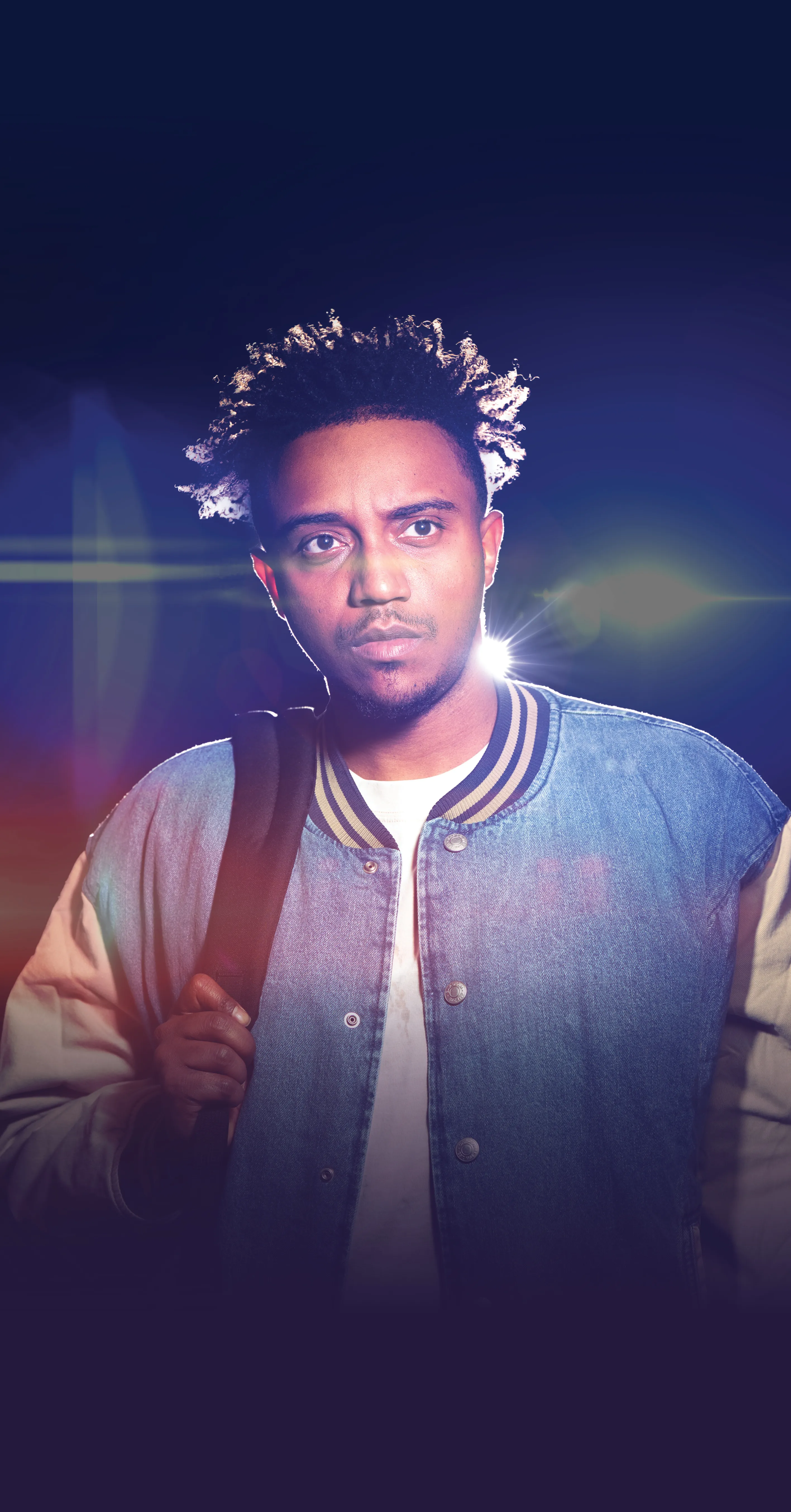 A man with short, spiked hair wears a denim varsity jacket and holds a strap over his shoulder, standing against a dark background with lens flare effects.