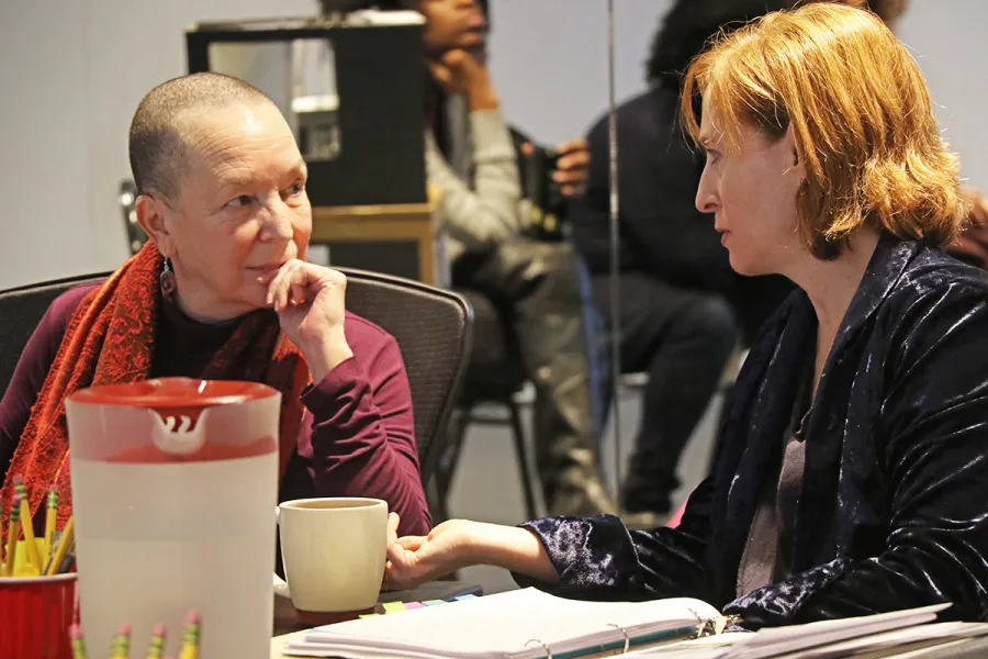 Two women sit at a table in conversation, one with a shaved head holding a mug, the other with shoulder-length hair; papers and a pitcher are on the table.
