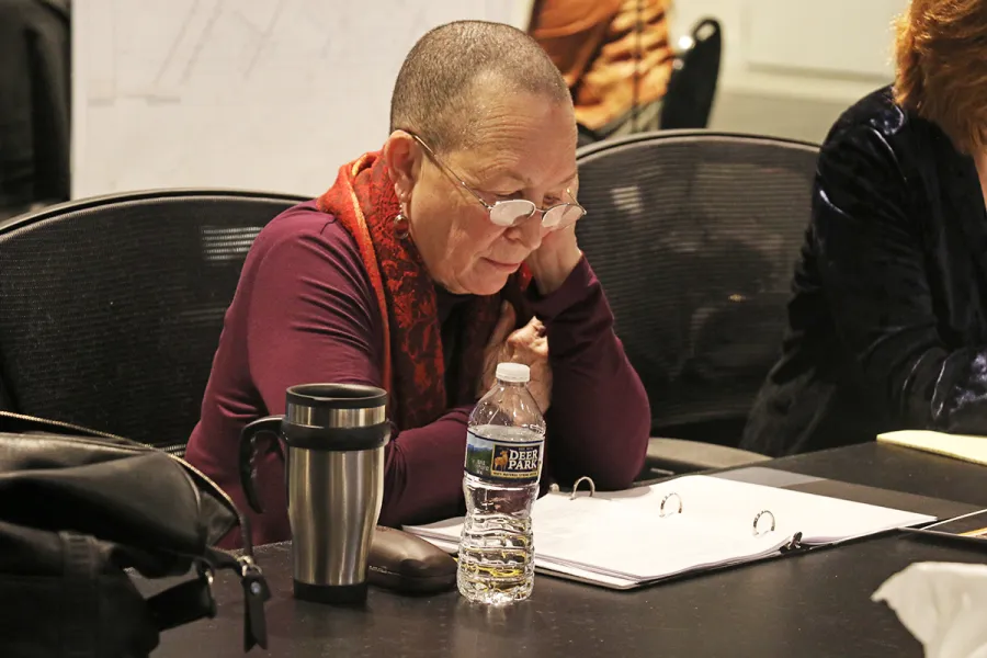 A person with short hair and glasses reads a binder at a table with a water bottle, travel mug, and bag nearby.