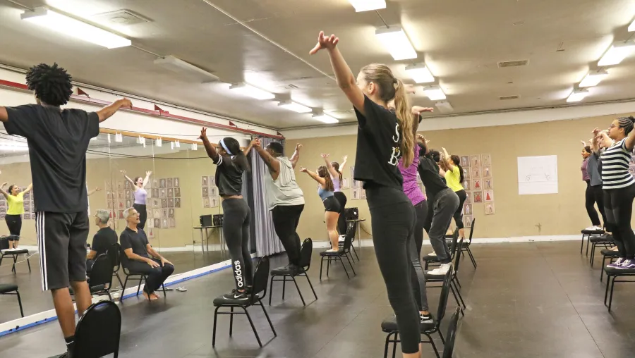A group of people in athletic wear stand on chairs in a dance studio, raising their arms, as two seated instructors observe.