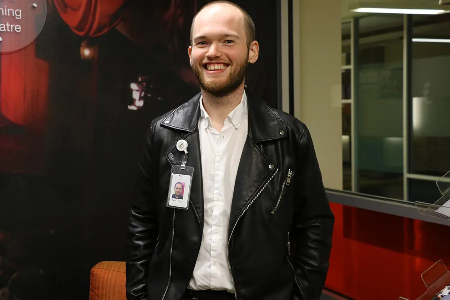 A man wearing a black leather jacket and a white shirt stands indoors, smiling, with an ID badge clipped to his jacket.