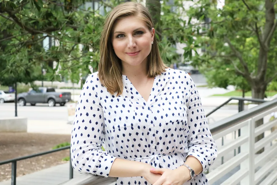 A woman with shoulder-length brown hair in a white dress with navy blue spots stands outdoors, leaning on a railing, with trees and cars in the background.