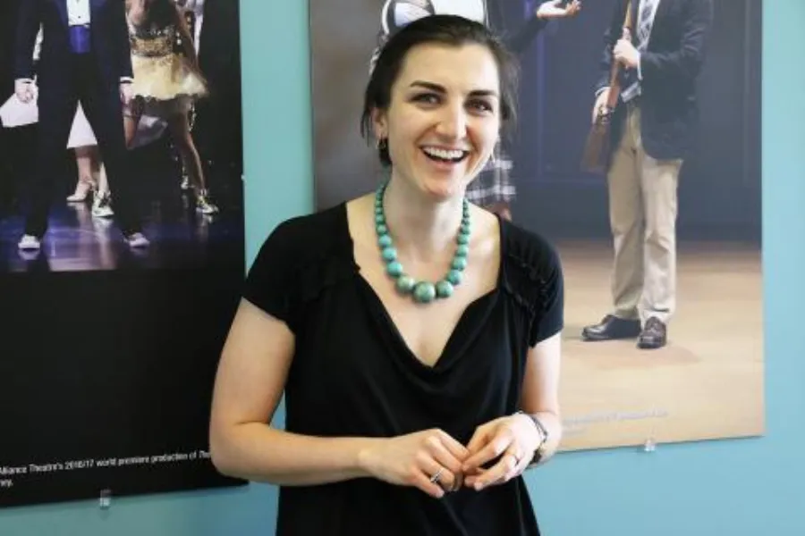 A woman wearing a black top and turquoise necklace stands smiling in front of posters featuring theatrical scenes.