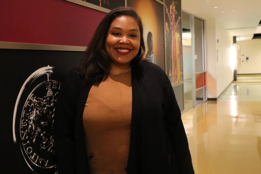 A woman with dark hair, wearing a brown top and black jacket, smiles while standing in a hallway with art displays on the walls.
