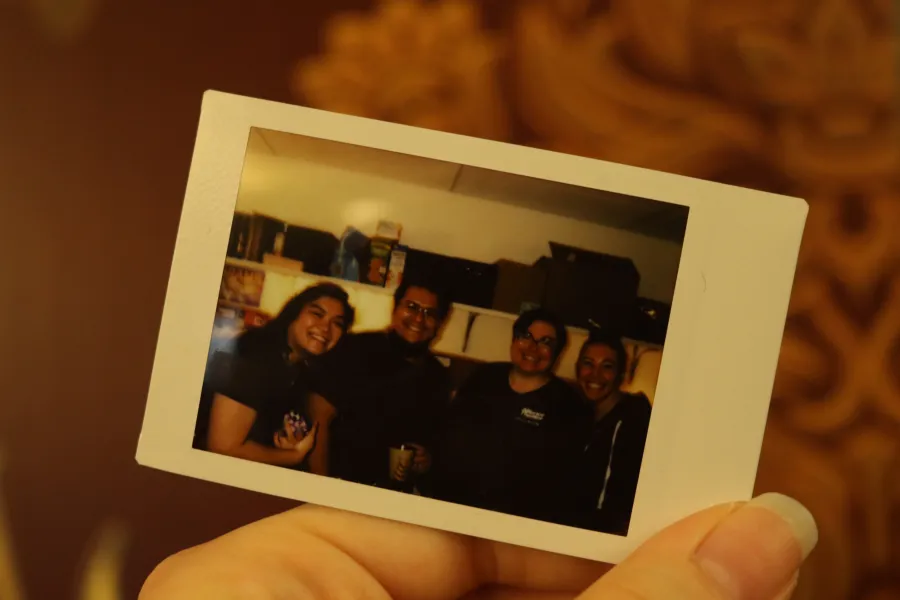 A hand holds a Polaroid photo showing four people standing together and smiling in a room with shelves in the background.