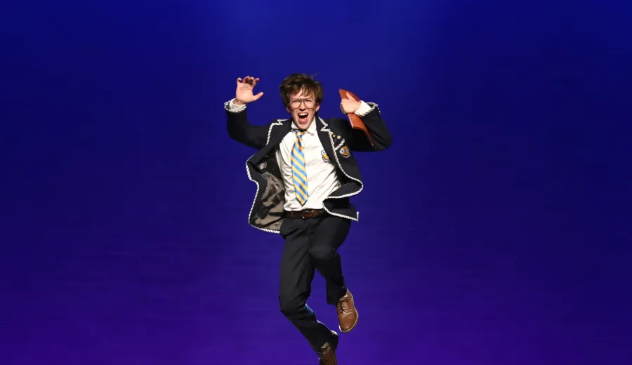 A person in a suit and tie jumps midair on stage against a solid blue background, raising their hands and making an animated facial expression.