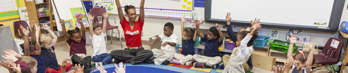A teacher sits on the floor with a group of young students in a classroom, all raising their hands in a circle during an activity.