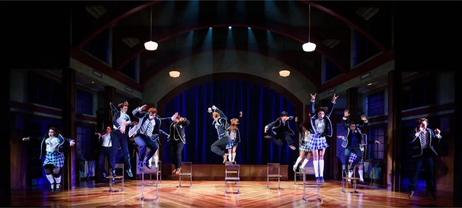 A group of performers in school uniforms jump in unison on stage, with dramatic lighting and a set resembling a school gymnasium in the background.