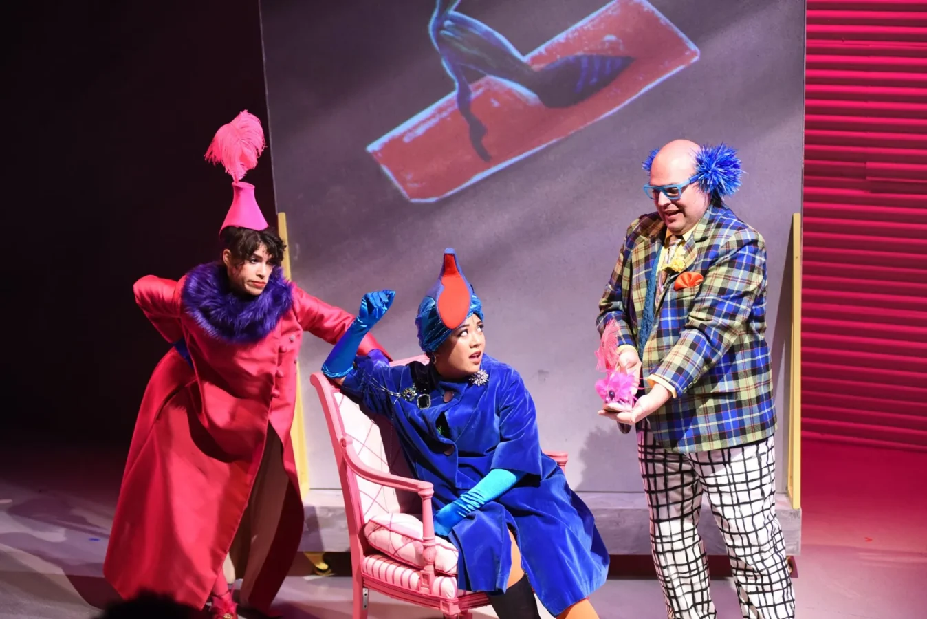 Three actors in colorful, whimsical costumes perform on stage, with one seated in a pink chair and another presenting a small pink object to the group.