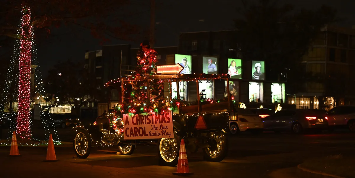 A vintage car decorated with Christmas lights and ornaments displays a sign reading "A Christmas Carol The Live Radio Play" in a parking lot at night.