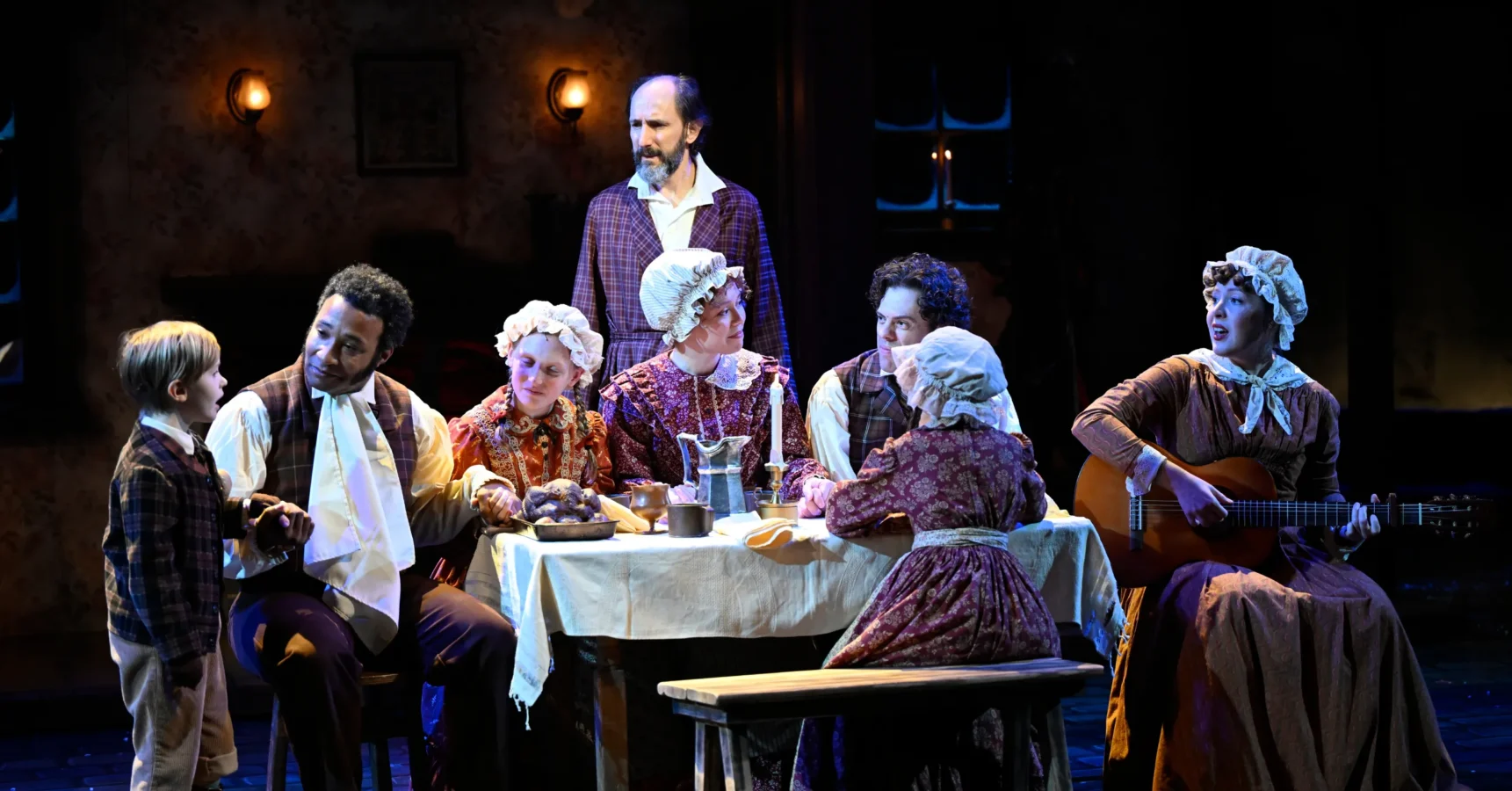A group of actors in period costumes perform a scene around a dining table on stage, with one playing a guitar and others engaging in conversation.