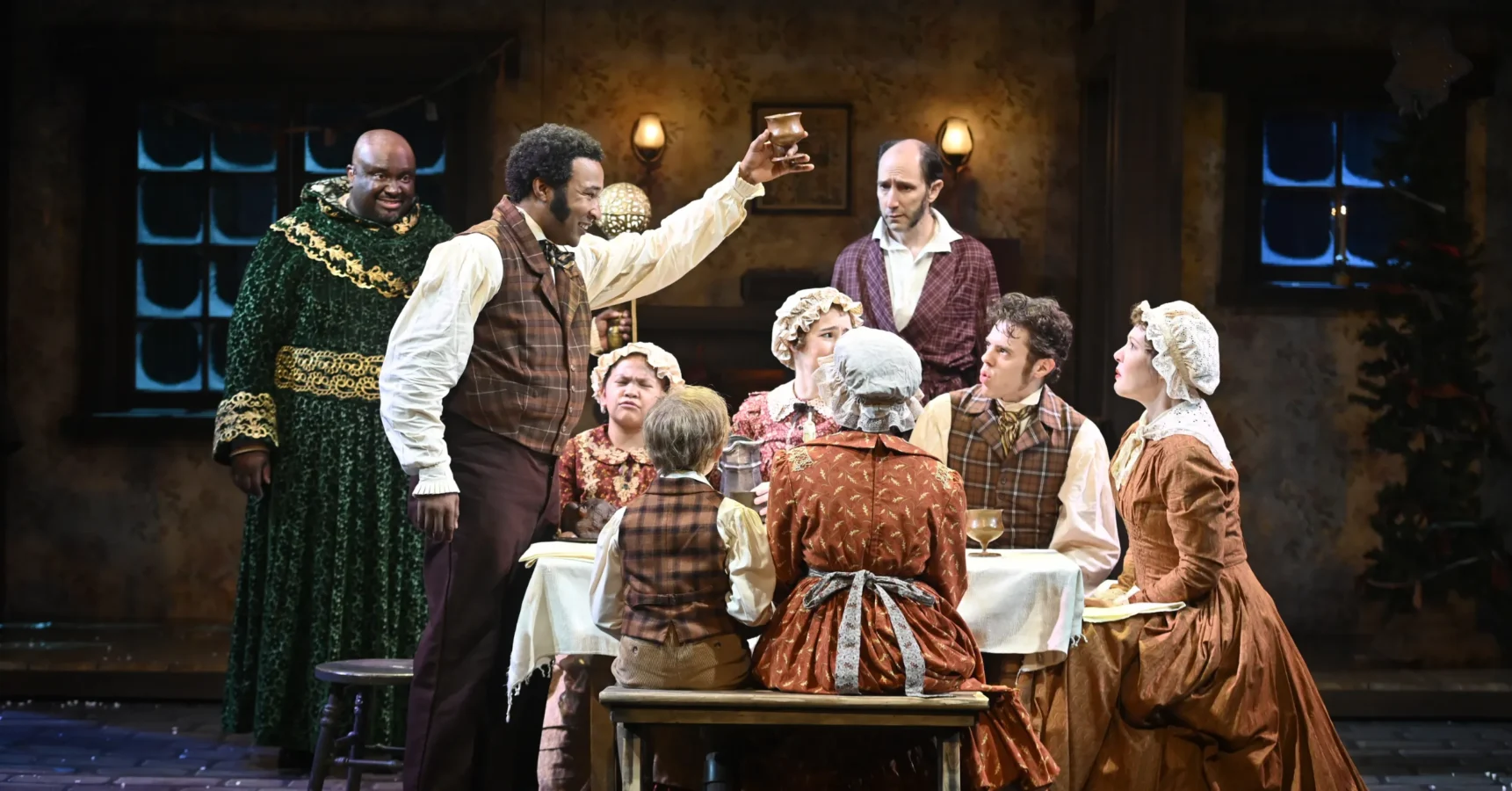 A group of actors in period costumes sits around a table on stage, with one man standing and raising a cup in a toast during a theatrical performance.