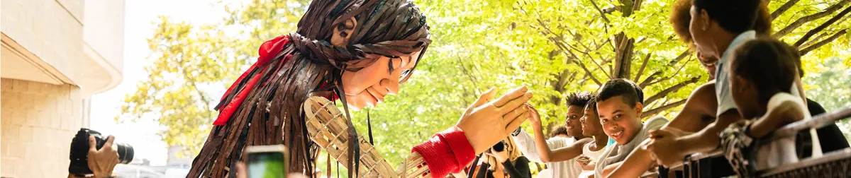 A large puppet figure with long brown hair and a red ribbon greets a group of smiling children and adults outdoors near a fence, with trees and sunlight in the background.