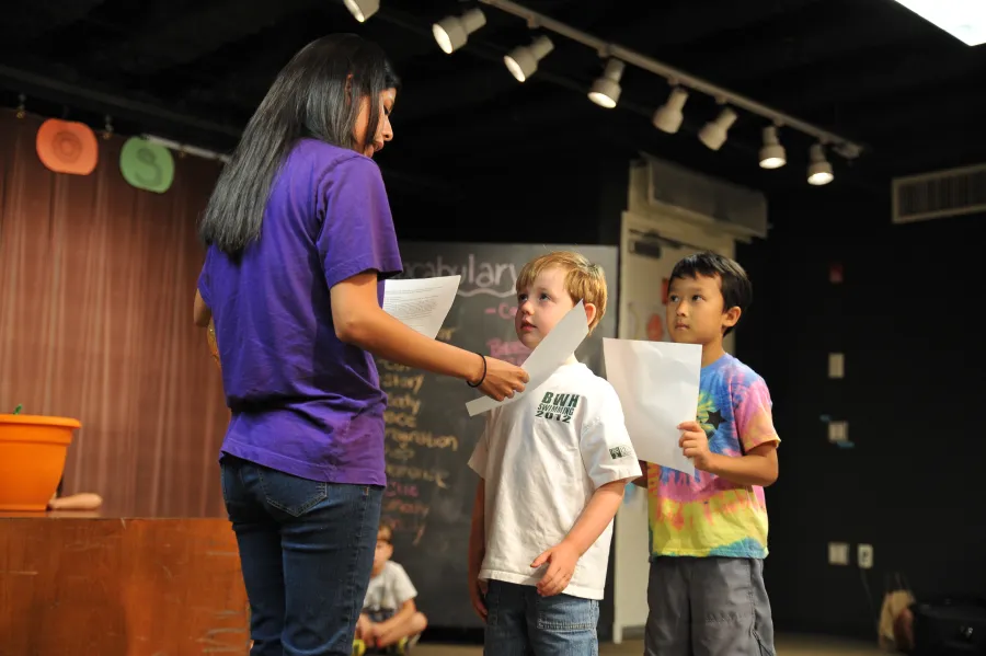 A woman hands papers to two young boys standing on a stage, with a brown curtain and a chalkboard in the background.
