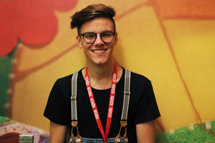 A young person with short brown hair, glasses, and a red lanyard smiles at the camera against a colorful, abstract background.