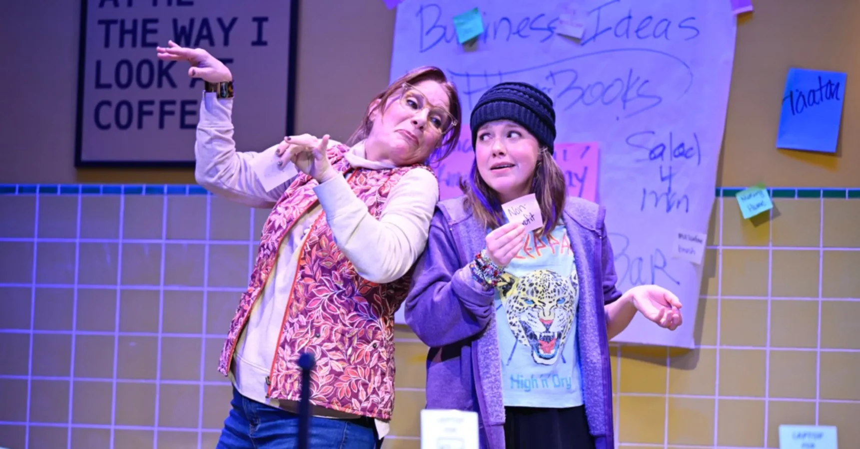 Two people stand in front of a tiled wall and a sign covered with notes. One leans back with raised arms, while the other holds up a piece of paper and wears a knit hat.