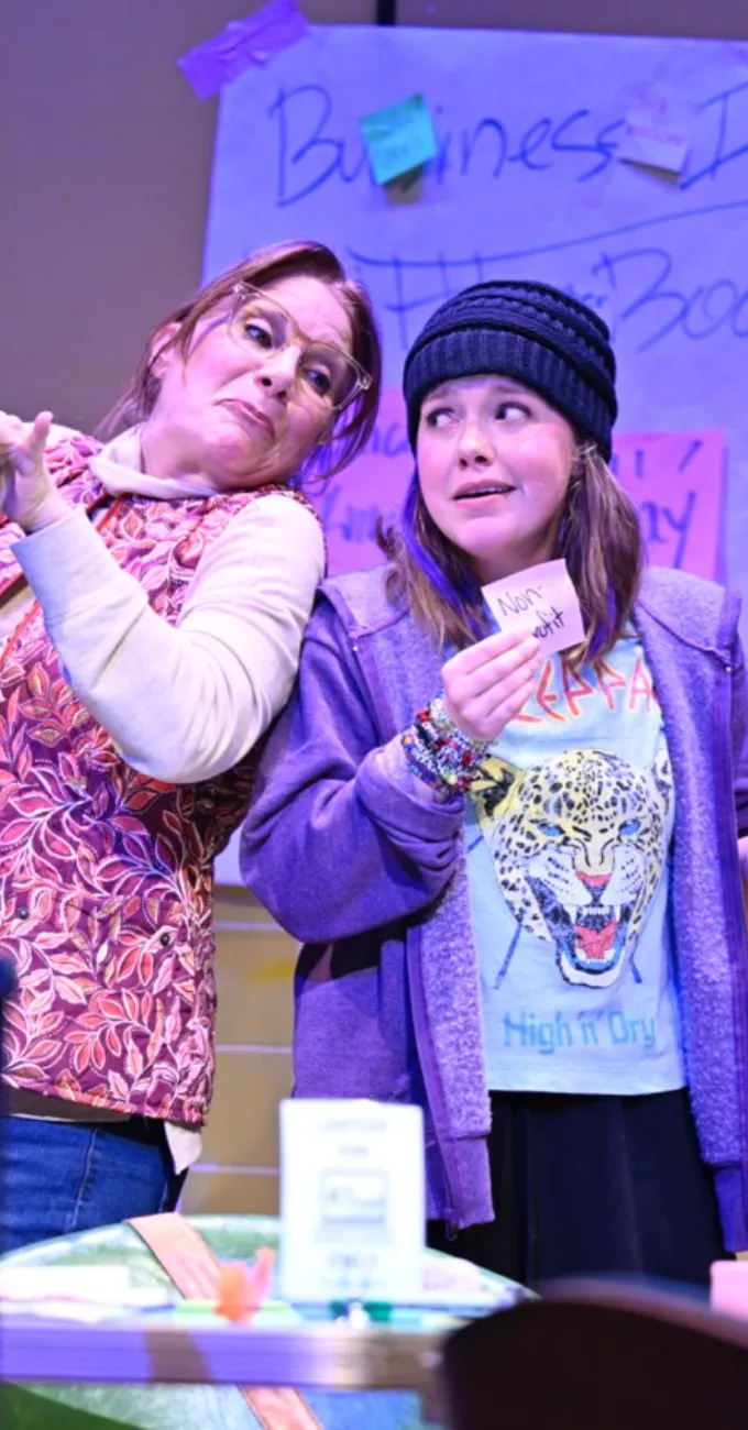 Two people stand side by side on stage; one holds a bat and looks surprised, while the other wears a hat, holds a note, and looks uncertain. A handwritten sign is visible in the background.