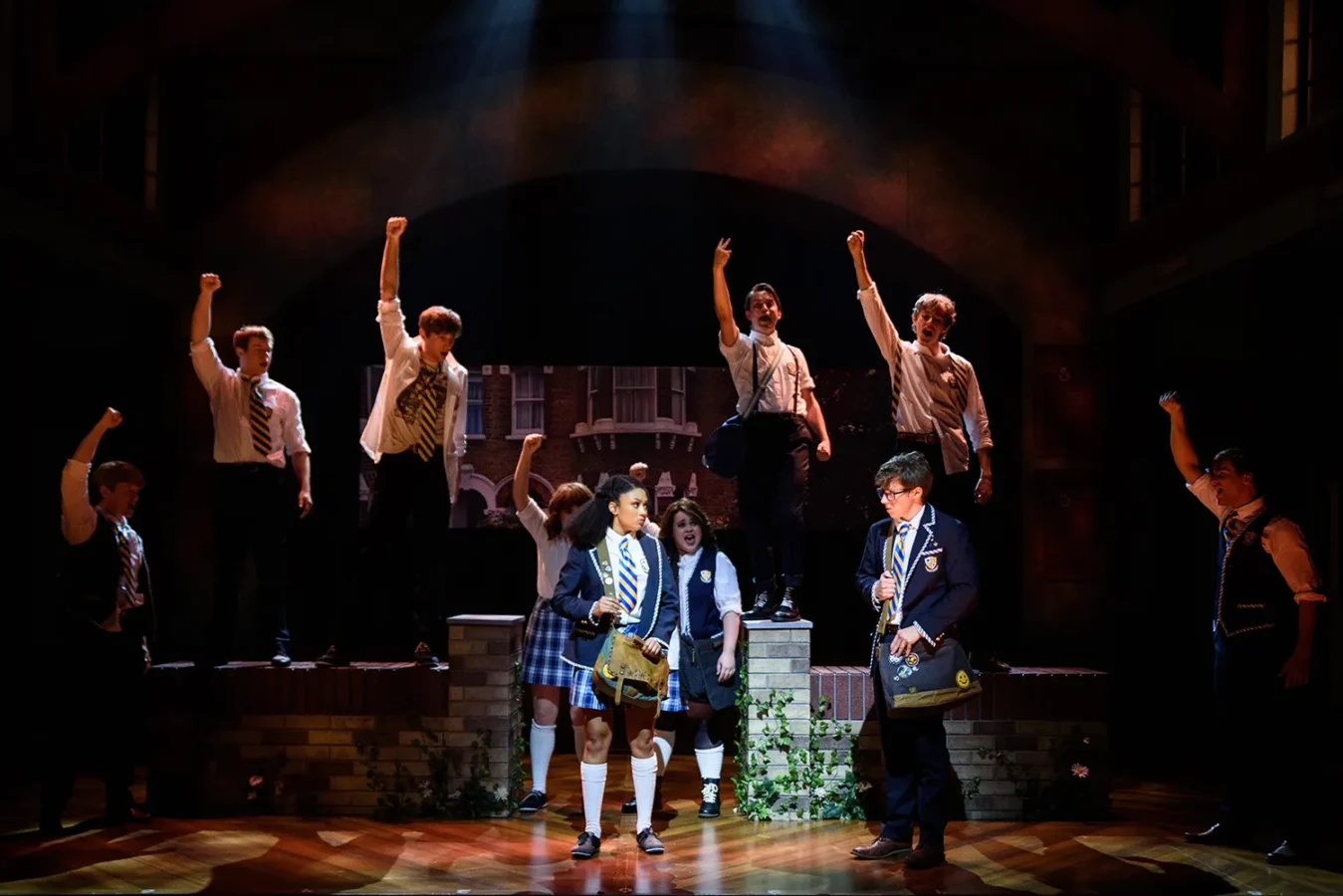 A group of students in school uniforms perform on stage, with several raising fists in the air under dramatic lighting.