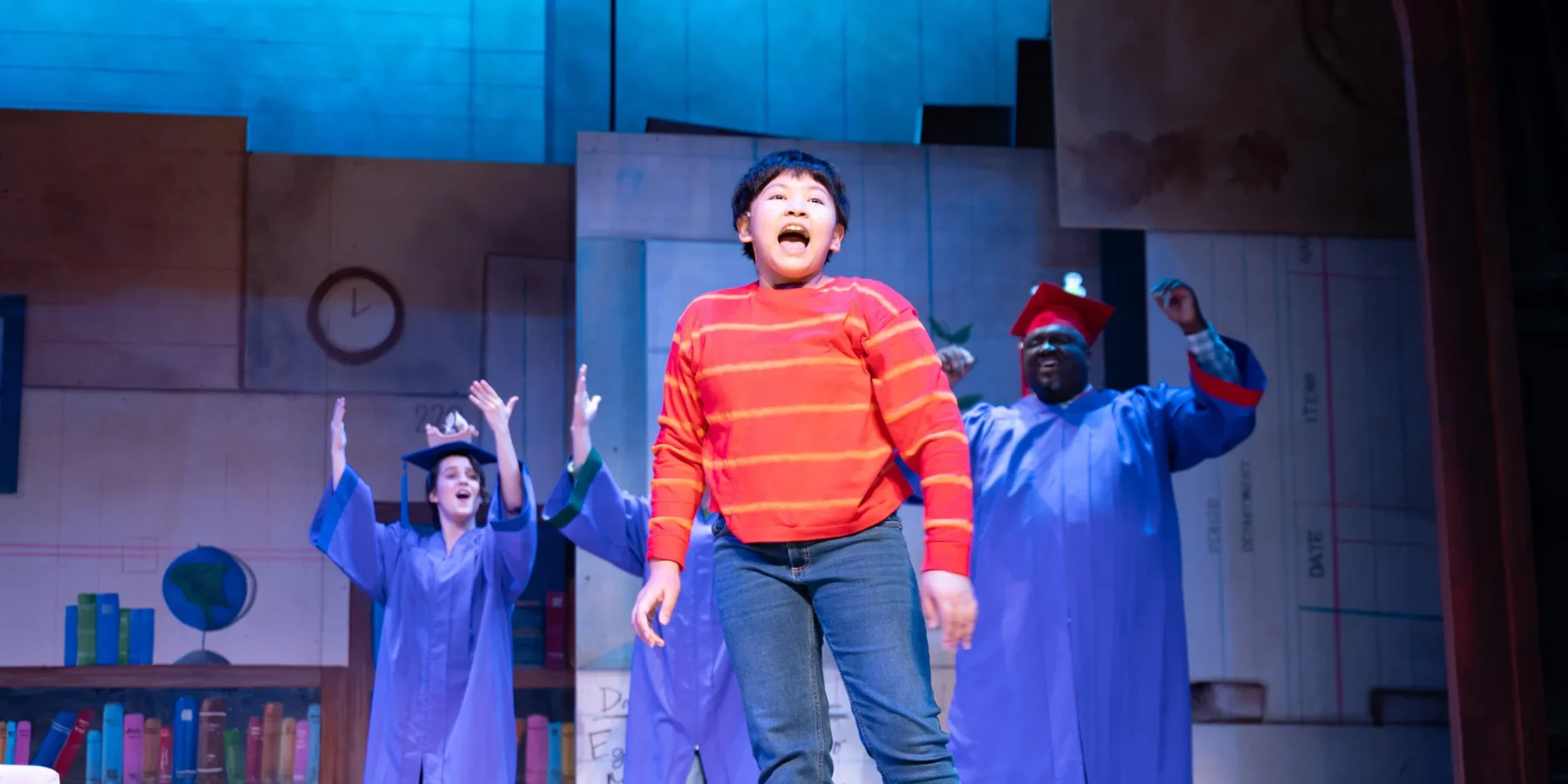 A child in a red striped shirt sings on stage in front of performers wearing blue graduation gowns and caps, with a blue-lit backdrop.