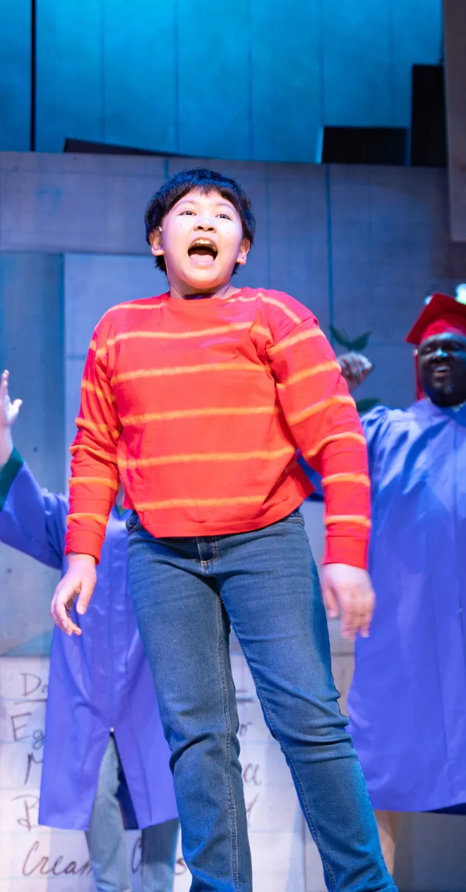 A child in a red and orange striped sweater and jeans performs on stage, with adults in blue gowns and caps standing in the background.