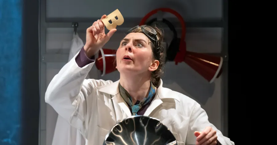 A person in a white lab coat and goggles holds up a small beige object, appearing to examine it closely, with lab equipment visible in the background.