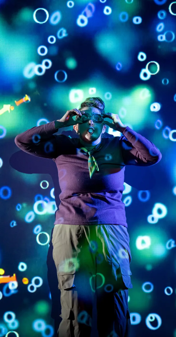 A person wearing glasses and a purple sweater stands in front of a screen displaying glowing blue and green bubbles or abstract circles.