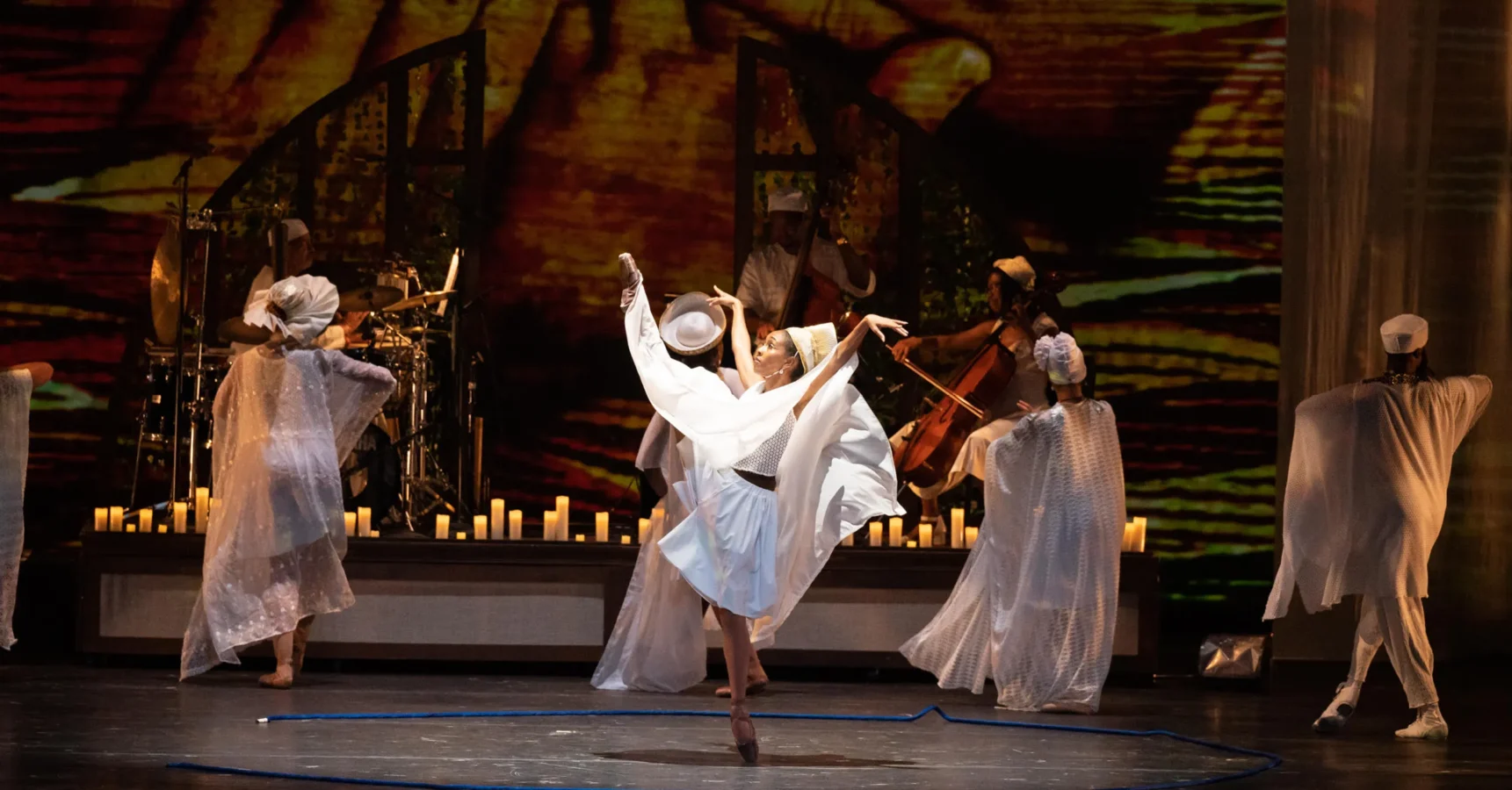 Dancers in flowing white costumes perform onstage, surrounded by musicians and candles, with a warm, abstract background projection.