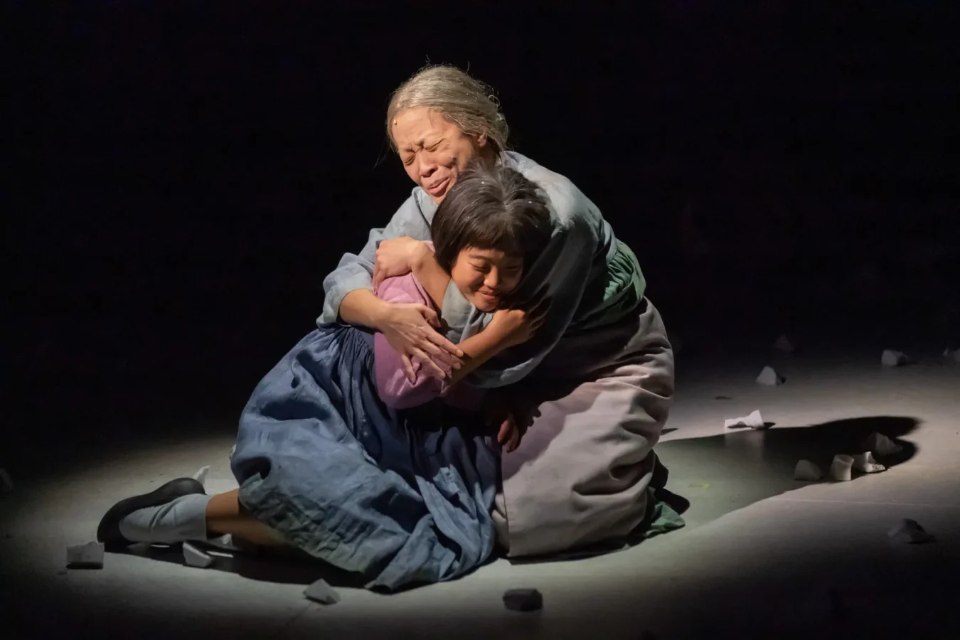 An older woman and a young girl sit on the floor in a spotlight, embracing tightly with emotional expressions, surrounded by scattered small white objects.