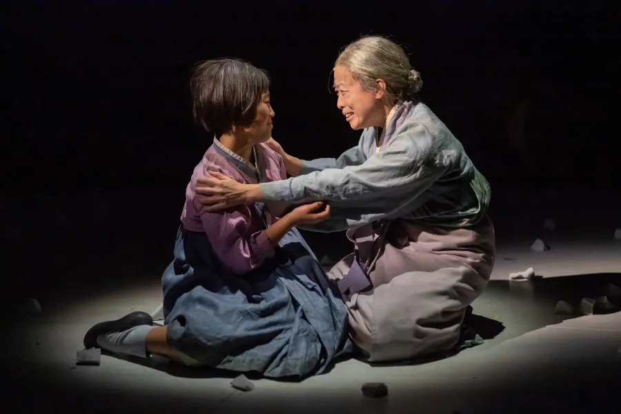 Two women in traditional Korean clothing sit on the floor, facing each other and embracing emotionally under a spotlight, with scattered white objects around them.