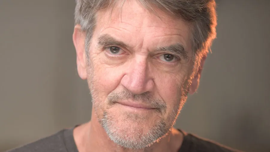 A middle-aged man with gray hair and a beard looks directly at the camera with a neutral expression, set against a plain, softly-lit background.